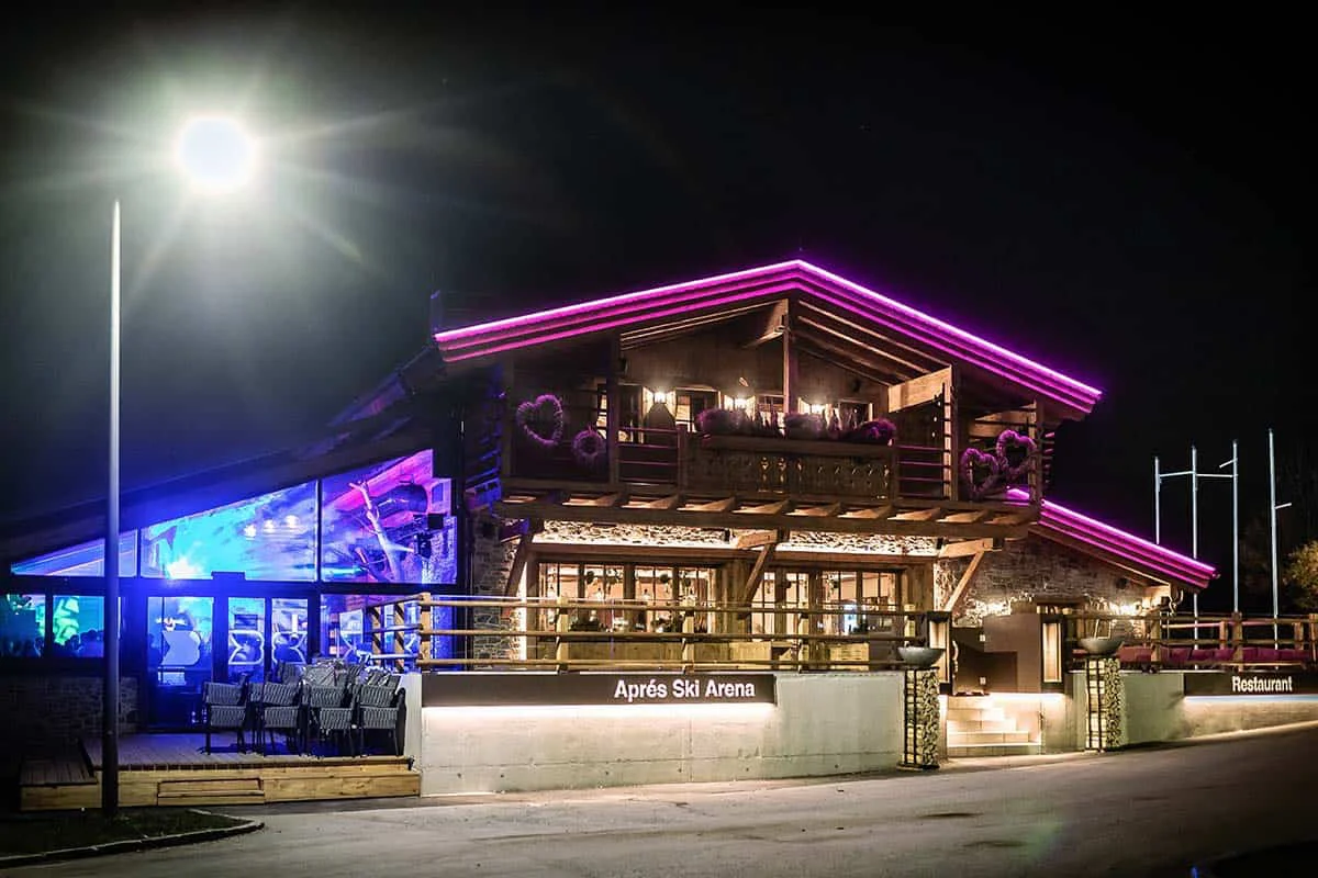 Holzhütte mit neonlila Beleuchtung und Terrasse, tagsüber geöffnet für Après-Ski und Restaurant.