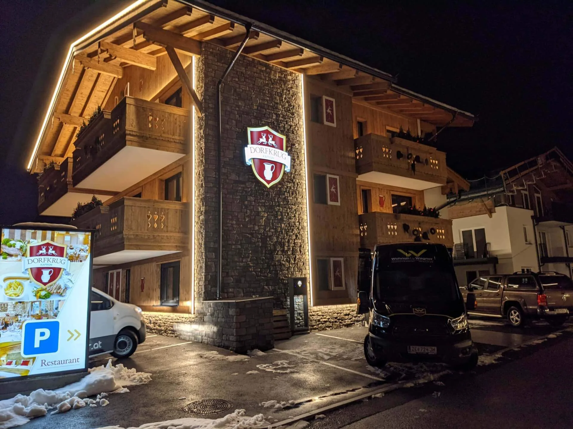 Dorfkrug-Gebäude mit beleuchtetem Schild und geparkten Fahrzeugen bei Nacht.
